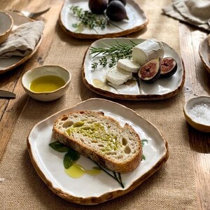 May include: A table setting with white ceramic plates and brown rims, displaying food. Sliced bread with olive oil, figs, cheese, and herbs are on the plates. Small bowls hold olive oil and salt. The setting has a rustic aesthetic.