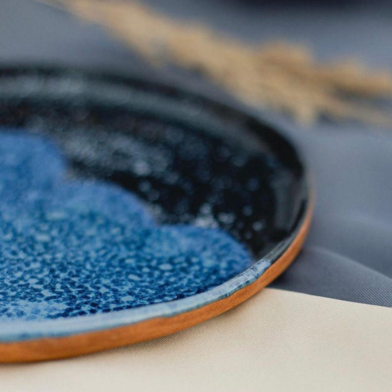 May include: A handmade ceramic plate with a unique design. The plate features a dark blue and light blue speckled glaze, with a natural brown rim. The plate is a perfect size for serving appetizers or small meals.