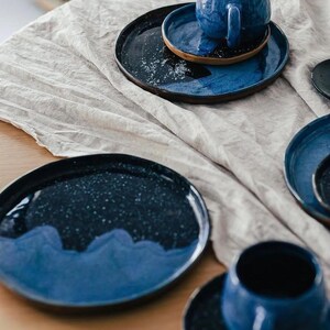 May include: A collection of handmade ceramic tableware in shades of blue. The set includes plates, saucers, and a mug, all with a speckled, glazed finish. The plates have a dark blue base with a lighter blue wave design.