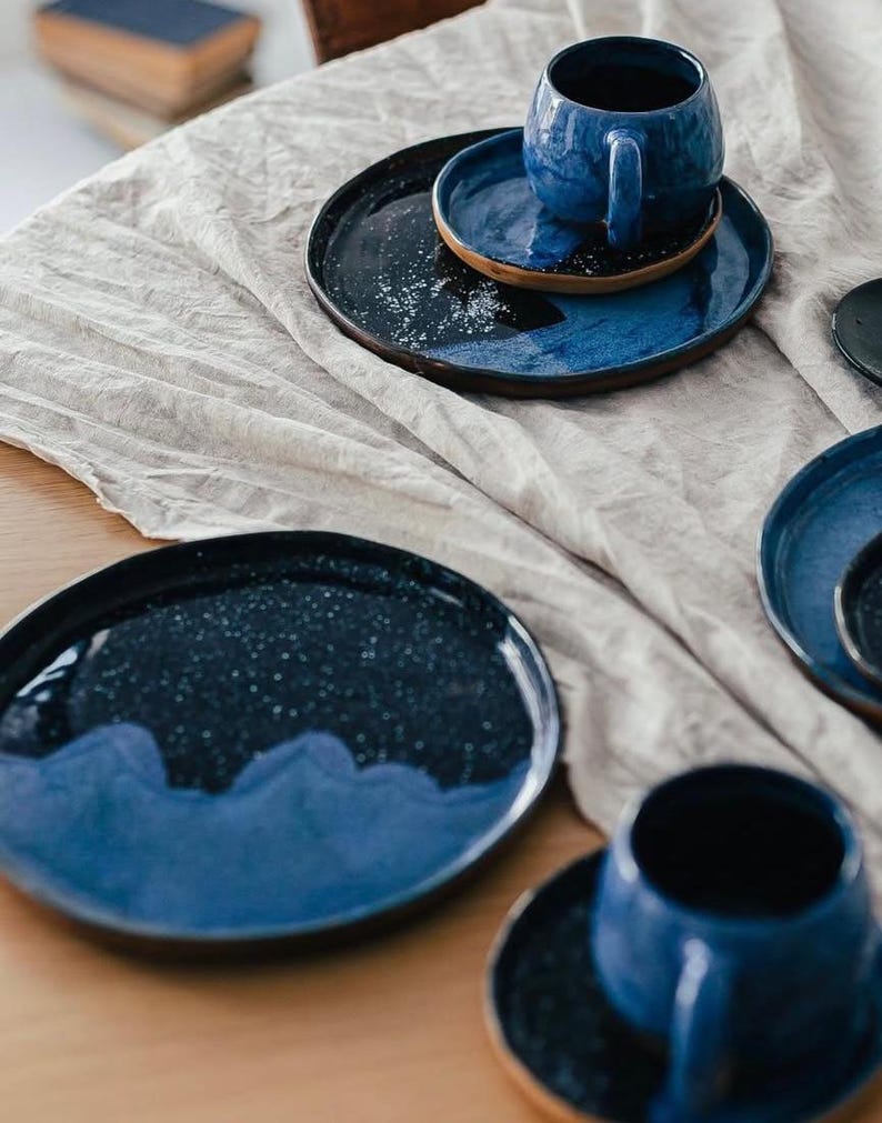May include: A collection of handmade ceramic tableware in shades of blue. The set includes plates, saucers, and a mug, all with a speckled, glazed finish. The plates have a dark blue base with a lighter blue wave design.