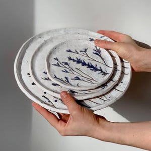 Puede incluir: Un juego de tres platos de cerámica hechos a mano. Los platos son blancos con un acabado moteado y presentan un delicado diseño floral azul. Los platos se sostienen frente a un fondo blanco.