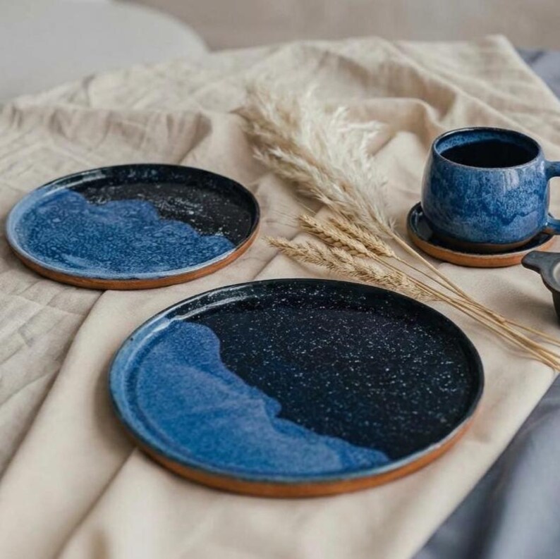 May include: Ceramic plates and a mug with a speckled blue and black glaze. The plates have a brown rim and a wave-like design. The mug sits on a matching saucer. A rustic, handmade aesthetic.