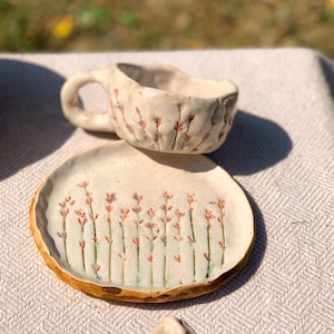 May include: A handmade ceramic cup and saucer set. The off-white pieces feature a floral design with pink and purple accents. The cup has a handle and the saucer has a slightly raised edge. The set is placed on a neutral-colored surface.