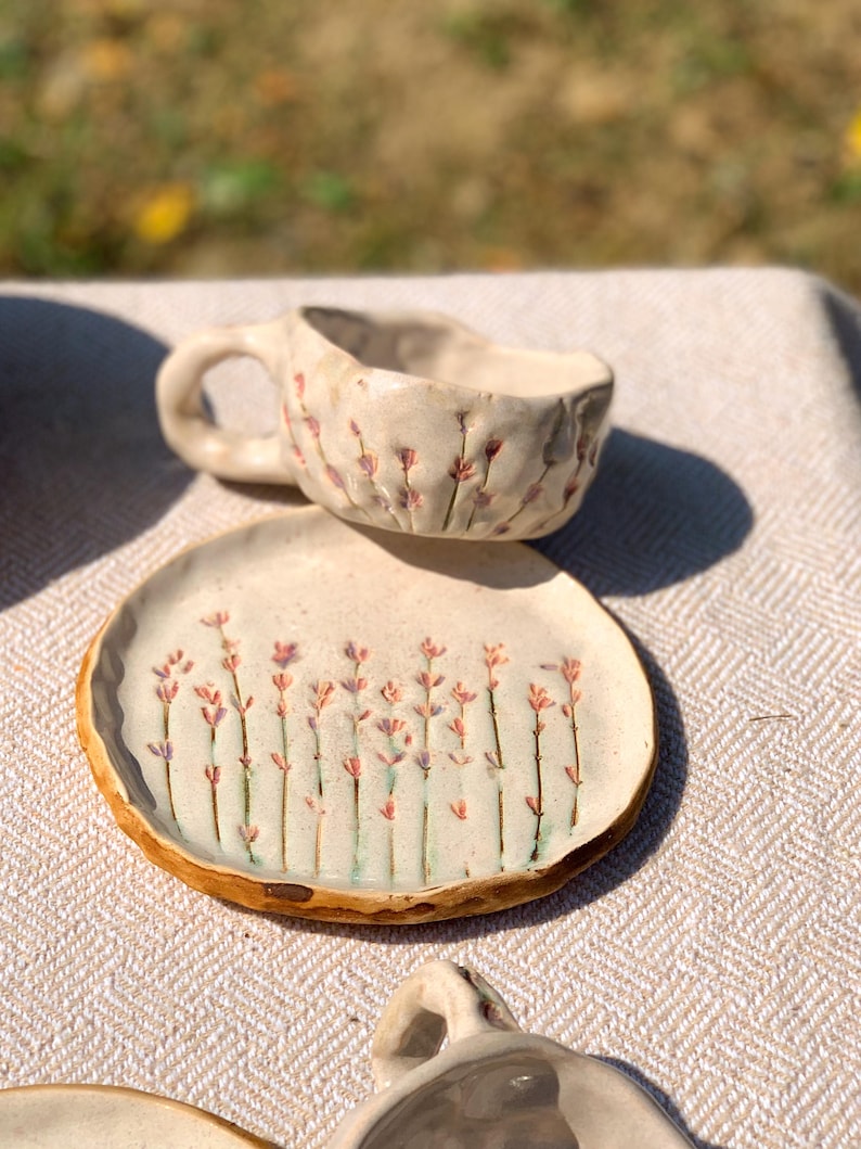 May include: A handmade ceramic cup and saucer set. The off-white pieces feature a floral design with pink and purple accents. The cup has a handle and the saucer has a slightly raised edge. The set is placed on a neutral-colored surface.