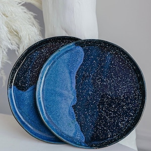 Puede incluir: Dos platos redondos de cerámica con un diseño moteado en azul oscuro y azul claro. Los platos tienen un acabado brillante y un borde oscuro. Los platos están apilados uno encima del otro.