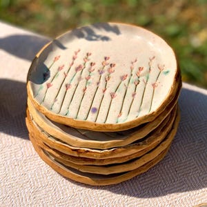 May include: A stack of five round ceramic plates with an off-white speckled glaze. Each plate features a hand-painted design of vertical stems with small pink, purple, and green floral accents. The edges of the plates are uneven and rustic.