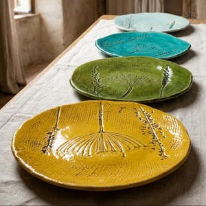 Pode incluir: Um conjunto de quatro pratos redondos de cerâmica dispostos em fila, com padrões botânicos em relevo. Os pratos são coloridos em amarelo, verde, azul-petróleo e azul claro, cada um com acabamento brilhante e detalhes texturizados de plantas.