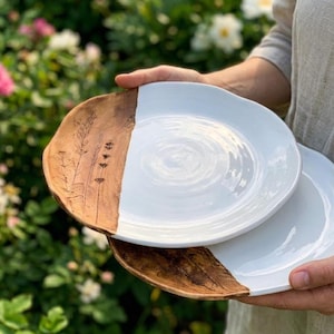 Op de afbeelding: Twee ronde keramische borden met een tweekleurig ontwerp. Eén kant van elk bord is afgewerkt met een glanzend wit glazuur, terwijl de andere kant een natuurlijke bruine kleitextuur toont met een botanische afdruk van hoge grassprieten.