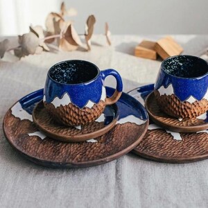 May include: A set of two ceramic mugs and matching plates. The mugs are blue with white mountain designs and brown textured bases. The plates are brown with white mountain designs. The mugs sit on smaller plates, and the set is on a neutral surface.