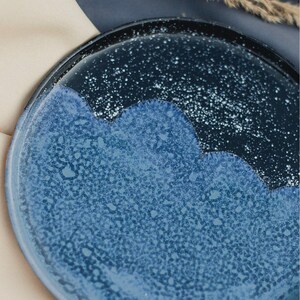 May include: A ceramic plate with a unique design. The plate features a dark blue rim that transitions into a lighter blue center with a speckled pattern. The edge of the plate has a wavy design, adding to its artistic appeal.
