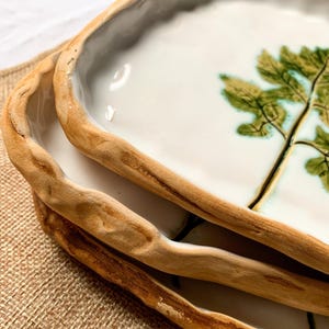 May include: Close-up of three ceramic serving dishes. Each dish has a white interior with a green and brown tree design. The rims are a light brown, textured, and organic shape. The dishes are stacked on top of each other.