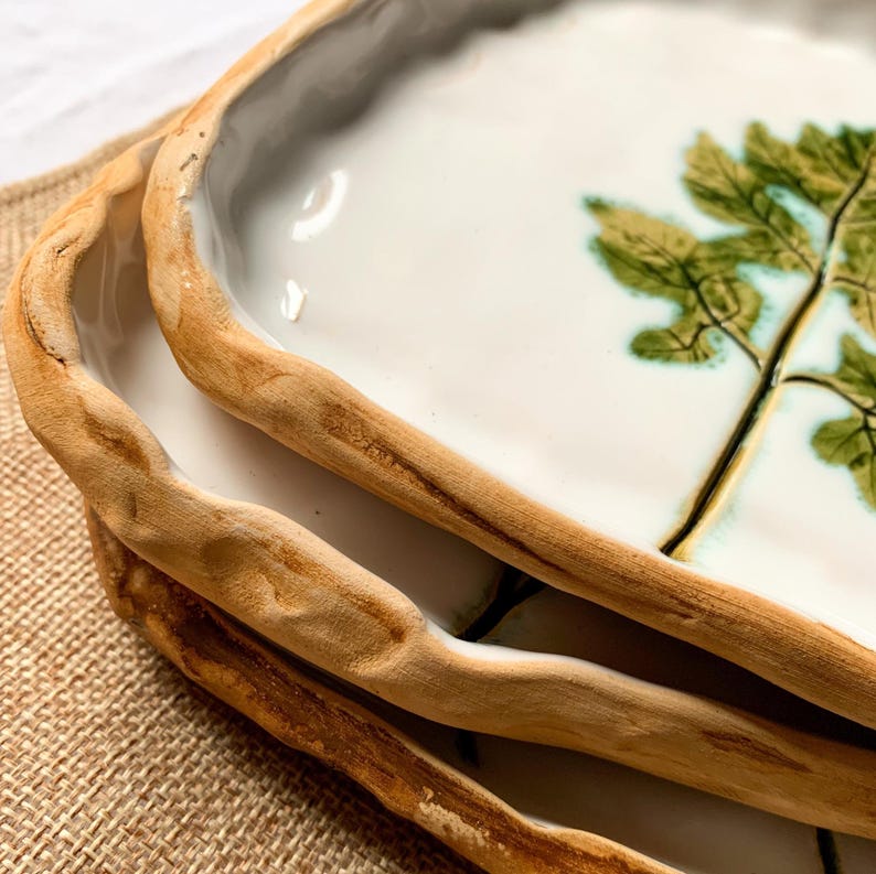 May include: Close-up of three ceramic serving dishes. Each dish has a white interior with a green and brown tree design. The rims are a light brown, textured, and organic shape. The dishes are stacked on top of each other.