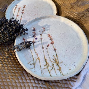 Pode incluir: Dois pratos de cerâmica branca com raminhos de lavanda e salpicos rosa. Os pratos têm uma borda ligeiramente levantada e estão colocados em papel de favo de mel castanho. Um ramo de lavanda seca está em primeiro plano.