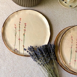 以下が含まれることがあります： 手描きのラベンダーのデザインと金色の縁が付いた、クリーム色のセラミックプレート。プレートには、ピンクとグリーンのアクセントが付いた花のモチーフがあります。乾燥したラベンダーの束がプレートの近くに置かれています。