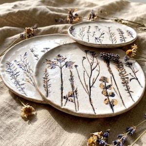 May include: Three round ceramic plates with an off-white speckled glaze. Each plate features dark, pressed botanical imprints of various wildflowers and grasses. The surface of the plates has a glossy finish, and the edges are slightly irregular.