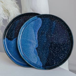 May include: Two ceramic plates with a speckled, dark blue and light blue design. The plates have a dark blue rim and a speckled pattern that resembles a starry night sky. The plates are stacked on top of each other.