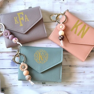 Pode incluir: Três pequenas carteiras personalizadas em tons de cinza, rosa e verde-azulado. Cada carteira apresenta uma inicial ou monograma dourado e um chaveiro decorativo com contas e detalhes florais. As carteiras são projetadas para guardar cartões e dinheiro.