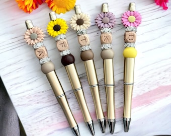 May include: Five gold-colored pens with decorative floral tops and lettered beads. Each pen features a different flower design: a daisy, a sunflower, and others in shades of pink, beige, and purple. The pens are arranged on a white and gray surface.