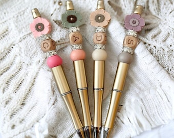 May include: Four gold pens with decorative tops. Each pen features a different colored flower, wooden beads with letters, and sparkling accents. The pens have a sleek, metallic finish and are arranged on a white, textured surface.