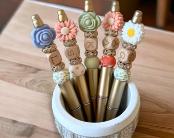 May include: Five decorative pens with gold-colored barrels and floral or beaded accents. Each pen features a different design, including a blue rose, pink daisy, green rose, pink flower, and white daisy. The pens are displayed in a white and gray textured holder.