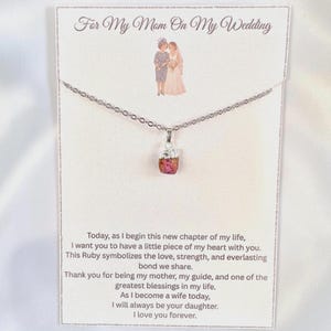 May include: A silver-toned necklace with a raw, reddish-pink ruby pendant on a display card. The card features an illustration of two people and the text: For My Mom On My Wedding, followed by a message about the ruby symbolizing love and strength.