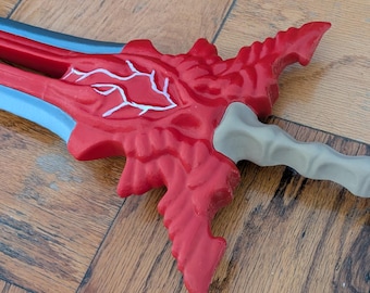 Peut inclure: Une épée fantastique rouge et bleue avec une garde rouge détaillée et une poignée gris clair. La garde présente un motif blanc en forme de veines. L'épée est posée sur une surface en bois.