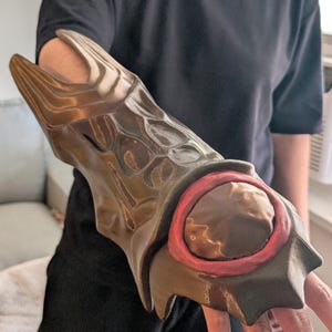 May include: A brown and bronze-colored arm guard with a textured, scale-like design. The guard features a circular brown element with a red border. The arm guard is worn on the arm.