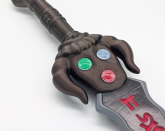 Peut inclure: Une épée jouet de couleur bronze foncé avec une poignée ornée de trois gemmes circulaires incrustées en vert, bleu et rouge. La garde présente un design incurvé en forme de corne. La lame est grise, texturée et porte des lettres rouges en relief.