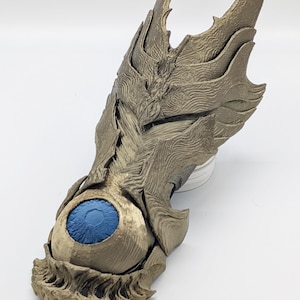 May include: A metallic gold gauntlet featuring a layered, textured armour design with sharp, pointed edges. A prominent, spherical blue eye with a dark pupil is embedded in the centre of the hand section, surrounded by intricate, wave-like patterns.