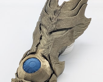 Peut inclure: Un gantelet en or métallique présentant un design d'armure texturé et superposé avec des bords pointus. Un œil bleu sphérique proéminent avec une pupille sombre est intégré au centre de la main, entouré de motifs complexes en forme de vagues.
