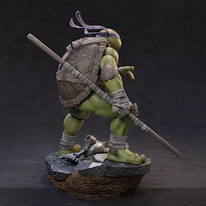 May include: A detailed figurine of Donatello from Teenage Mutant Ninja Turtles. The figure is green with a brown shell and purple head wrap. He holds a bo staff, standing on a rocky base. The figure is a collectible item.