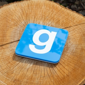 May include: Square blue coaster with a white "g" logo. The coaster has a white border and is set on a wooden surface. The logo is a stylized lowercase letter "g".