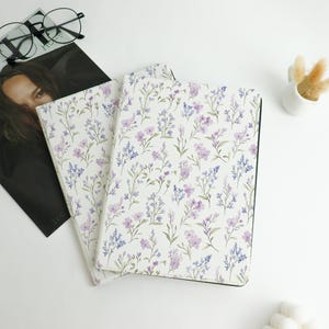 May include: Two notebooks with floral patterns. The covers feature a white background with a repeating design of purple and blue flowers and green stems. The notebooks are stacked on top of each other, with a magazine and glasses in the background.