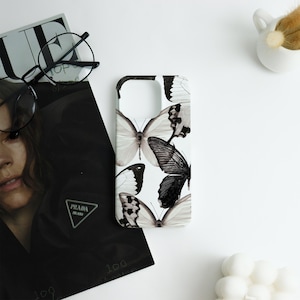 May include: A white phone case featuring a pattern of black and white butterflies. The butterflies have detailed wing patterns. The case is on a white surface next to a magazine, a pair of glasses, and a small white vase.