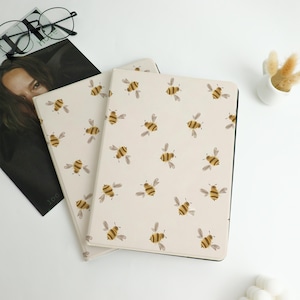 May include: Two cream-coloured notebooks with a repeating pattern of cartoon bees. The notebooks are open, revealing black edges. A magazine, glasses, and a small vase with dried flowers are in the background.