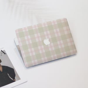 Könnte beinhalten: Ein Laptop mit einem pastellrosa und grünen Karomuster. Der Laptop hat ein weißes Apple-Logo in der Mitte. Er liegt auf einer weißen Oberfläche, mit einer Zeitschrift in der linken unteren Ecke.