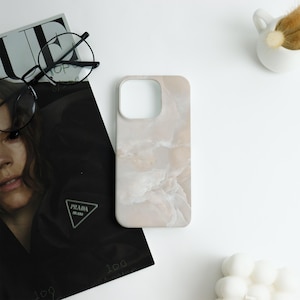 May include: A light beige phone case with a marble-like pattern. The case is lying on a white surface next to a black magazine, a pair of glasses, and a small white decorative item.