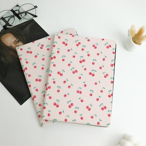 May include: Two notebooks with a light pink cover featuring a pattern of red cherries with green stems. The notebooks are lying on a white surface, with a magazine and glasses in the background. The notebooks have black edges.