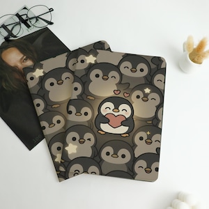 May include: Two notebooks with a repeating pattern of cartoon penguins in shades of gray and black. One penguin holds a pink heart. The notebooks are on a white surface with a magazine and glasses in the background.