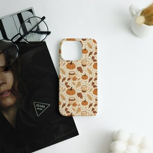 May include: A beige phone case featuring a fall-themed pattern. The design includes orange pumpkins, brown leaves, and coffee cups. The case is surrounded by a magazine, glasses, and a small white decorative item.
