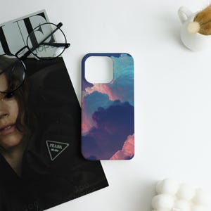 May include: A smartphone case with a vibrant cloud design in shades of blue, pink, and purple. The case has a dark blue border and a cutout for the camera. The case is lying on a white surface next to a magazine and a pair of glasses.