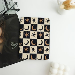 May include: A rectangular notebook with a celestial pattern of stars and crescent moons in a checkerboard design. The alternating squares are black and cream-colored. The stars and moons are a bronze color. The notebook is on a white surface.