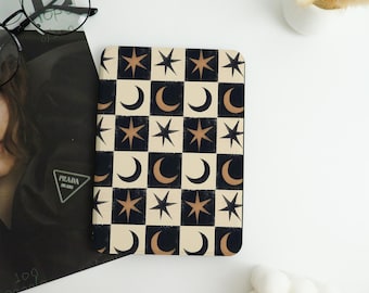 May include: A rectangular notebook featuring a celestial pattern of stars and crescent moons in a checkerboard design. The alternating squares are black and cream. The stars and moons are bronze. The notebook is on a white surface.