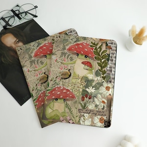 Könnte beinhalten: Drei Notizbücher mit einer skurrilen Illustration eines Frosches mit roter Pilzkappe, umgeben von Blumen und Blättern. Die Notizbücher haben eine Vintage-Ästhetik mit Text- und Schmetterlingsdetails. Der Satz "you drew stars around my scars" ist sichtbar.
