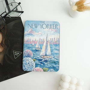 Könnte beinhalten: Ein rechteckiger Untersetzer mit einer Aquarellillustration von Segelbooten auf blauem Meer und einer Skyline. Der Untersetzer trägt den Text "THE NEW YORKER" oben und ist von blauen und rosa Blumen umgeben.