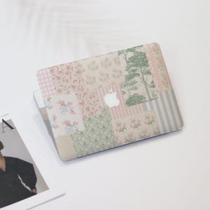 May include: A laptop case with a patchwork design featuring floral, striped, and tree patterns in pastel colors. The case has a white Apple logo in the center. The case is on a white surface.