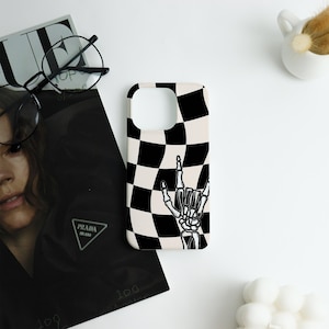 May include: A phone case featuring a black and white chequered pattern and a skeleton hand in a rock and roll pose. The case has a white base with a black and white chequered design. The skeleton hand is black and white.