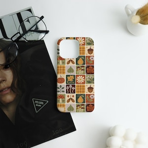May include: A phone case with a patchwork design featuring autumn-themed illustrations. The case displays squares with images of pumpkins, leaves, mushrooms, pears, and acorns in shades of orange, green, and brown. The case is on a white surface.