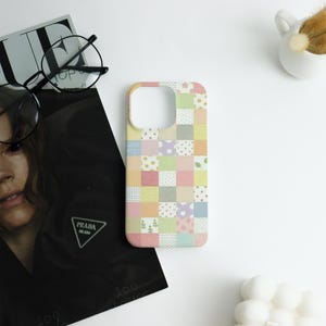 May include: A pastel-colored phone case with a patchwork design. The case features squares of various colors and patterns, including polka dots, floral prints, and geometric shapes. The text "GOOD BYE!" is visible on the case.