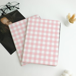 May include: Two notebooks with pink and white gingham pattern covers. The notebooks are stacked on top of each other, with the top one slightly angled. A magazine and eyeglasses are in the background.